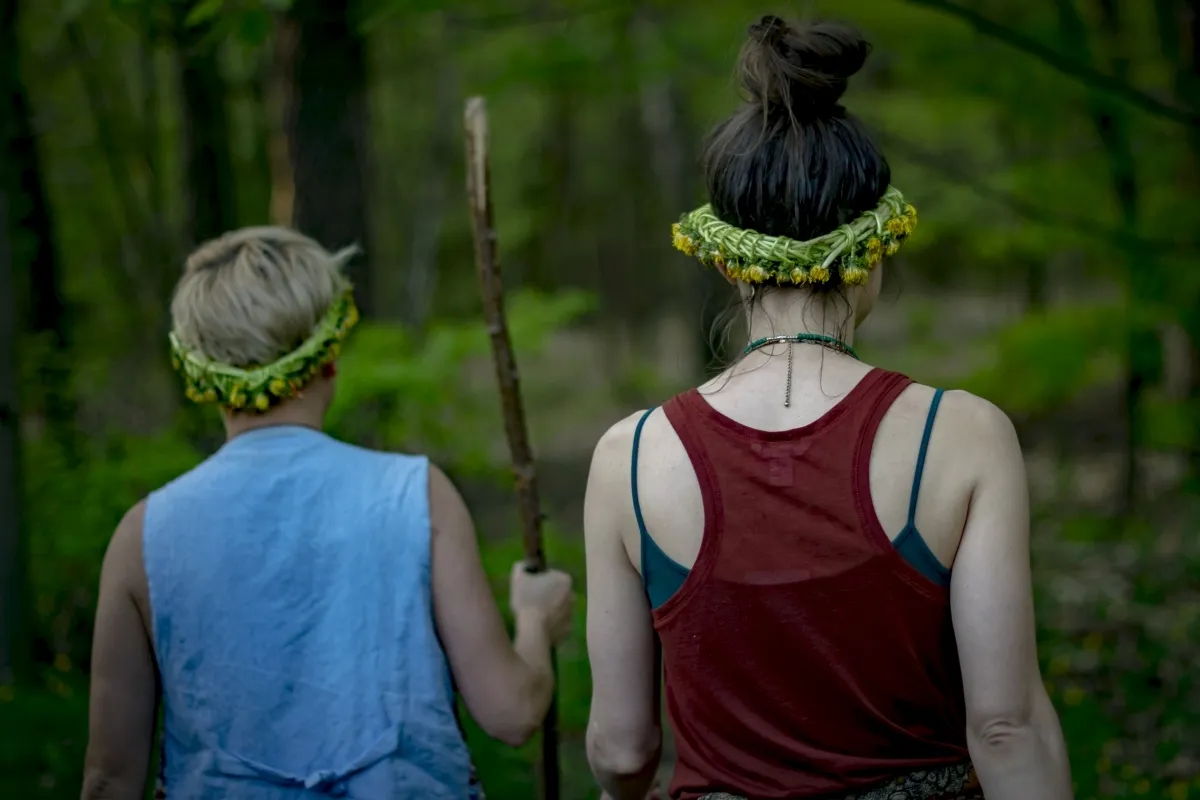 Two people wearing flower wreaths walking away through a green forest, one holding a stick